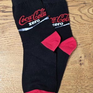 Könnte beinhalten: Paar schwarze Socken mit roten Fersen und dem Coca-Cola Zero-Logo in Rot und Weiß. Die Socken werden auf einer Holzoberfläche präsentiert.