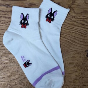 May include: White ankle socks featuring a black cat design with purple ears and a red bow tie. The socks have a purple stripe near the ankle and the word "JIJI" printed on the side. The cat design is repeated on the top of the socks.