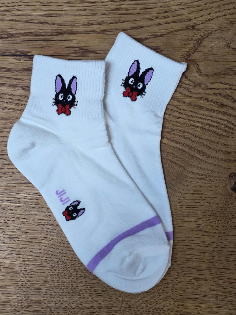 May include: White ankle socks featuring a black cat design with purple ears and a red bow tie. The socks have a purple stripe near the ankle and the word "JIJI" printed on the side. The cat design is repeated on the top of the socks.