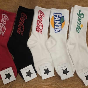 May include: Five pairs of crew socks with beverage brand logos. The socks are red, black, and white. The logos include Coca-Cola, Fanta, and Sprite. Each pair has a tag with a black star.