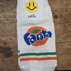 May include: White ankle socks featuring the blue and orange Fanta logo with a green leaf graphic. The socks have horizontal green and orange stripes near the cuff and include a white tag with a yellow smiley face and the word smile.