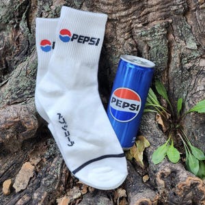 May include: A pair of white socks with a black Pepsi logo and Japanese text. A blue can of Pepsi is next to the socks.