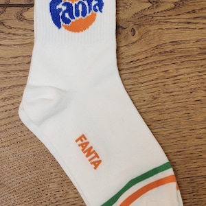 May include: White socks with a bright orange and blue Fanta logo and a green and orange stripe at the top. The socks have the word "Fanta" written in orange on the side.
