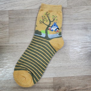 May include: Mustard yellow sock with a green and yellow striped pattern. The top of the sock features a cartoon illustration of a blue creature and two children sitting in a tree. The sock is made of a soft, comfortable material.