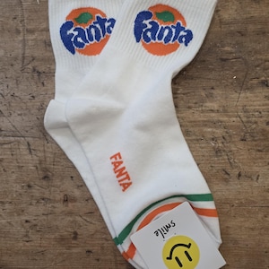 May include: A pair of white crew socks featuring the blue and orange Fanta logo on the cuff and the word Fanta in orange on the side. The socks have orange and green stripes near the toe and include a white tag with a yellow smiley face and the word smile.