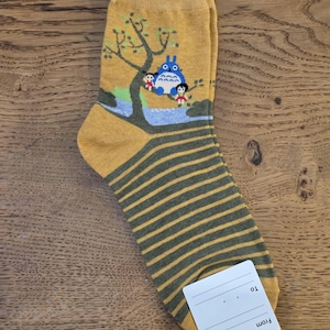 May include: Mustard yellow socks with a cartoon illustration of a blue creature and two children under a tree. The lower portion of the sock features horizontal olive green stripes. A white tag is attached.