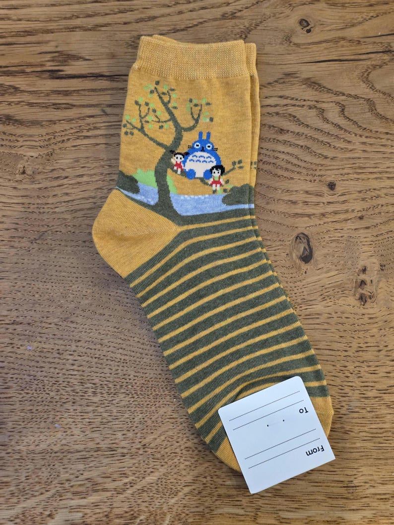 May include: Mustard yellow socks with a cartoon illustration of a blue creature and two children under a tree. The lower portion of the sock features horizontal olive green stripes. A white tag is attached.