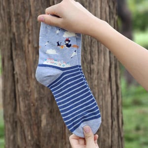 May include: A pair of blue and white striped socks with a cartoon image of a witch flying on a broomstick. The socks have a light blue background with white clouds and a small town.