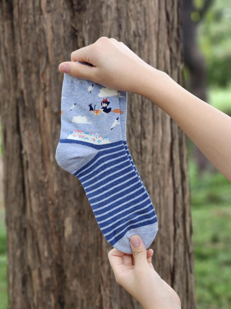 May include: A pair of blue and white striped socks with a cartoon image of a witch flying on a broomstick. The socks have a light blue background with white clouds and a small town.