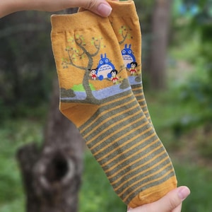 May include: Mustard yellow socks featuring a cartoon illustration of a blue creature, children, and a tree. The socks have grey and olive green horizontal stripes on the lower part. The socks are held up by a person.