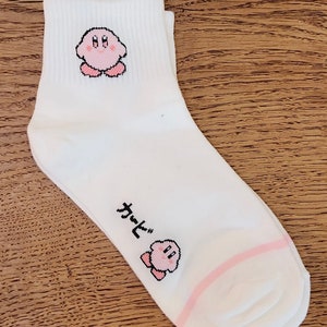 Peut inclure: Chaussettes blanches à la cheville avec un personnage de dessin animé rose. Le personnage est une créature rose avec un grand sourire et de grands yeux. Les chaussettes ont une bande rose autour du haut.