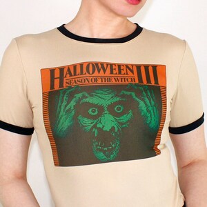 May include: A beige t-shirt with a black ringer and a graphic print of the "Halloween III: Season of the Witch" movie poster. The poster features a green and orange monster with a menacing expression.