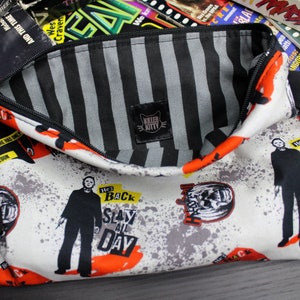 Pode incluir: Uma bolsa branca com zíper com um forro listrado preto e branco. A bolsa tem uma alça preta com fecho de pressão. A parte frontal da bolsa apresenta uma impressão gráfica de um personagem do filme "Halloween" com o texto "He's Back Slay All Day".