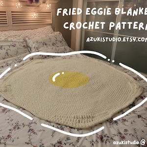 Könnte beinhalten: Eine weiße gehäkelte Decke in Form eines Spiegeleis mit einem gelben Eigelb. Die Decke liegt auf einem Bett mit einer Bettwäsche mit Blumenmuster. Der Text "FRIED EGGIE BLANKET CROCHET PATTERN AZUKISTUDIO.ETSY.COM" befindet sich oben im Bild.