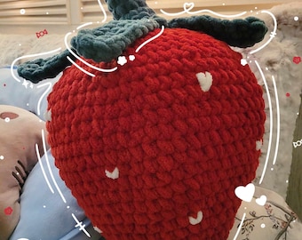 Peut inclure: Un coussin en forme de fraise rouge crocheté avec des accents blancs. Le coussin est sur une surface à motifs floraux blancs.