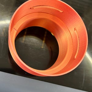 Può includere: Oggetto cilindrico arancione stampato in 3D con apertura circolare. L'oggetto presenta una serie di intagli curvi sul lato. L'oggetto è appoggiato su una superficie scura.