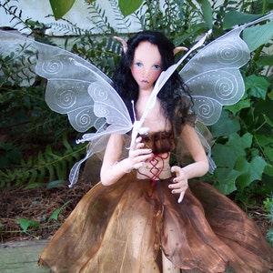 Könnte beinhalten: Eine handgefertigte Puppe mit langen dunklen Haaren, die ein braunes und goldenes Kleid mit einem weißen Tüllüberzug trägt. Die Puppe hält einen weißen Schmetterling mit durchsichtigen Flügeln. Die Puppe steht vor einem grünen, belaubten Hintergrund.