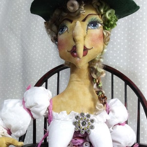 Pode incluir: Uma boneca de bruxa com um chapéu verde, um vestido branco e uma saia rosa está sentada em uma cadeira de madeira. Ela tem um nariz grande e olhos azuis. Ela está usando um broche de joias e uma fita dourada.