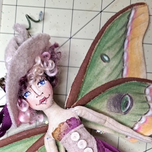 May include: A fabric doll featuring a wide-brimmed hat, curly pink hair, and blue eyes. The doll has large green moth wings with yellow and brown accents, a purple bodice with three white buttons, and black leggings with colorful polka dots.