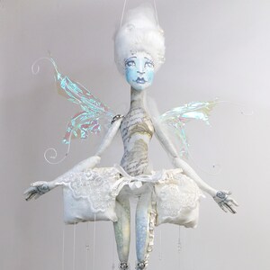 May include: A white fairy doll with iridescent wings, a white lace skirt, and blue and white patterned legs. The doll is hanging from the ceiling with strings of beads.