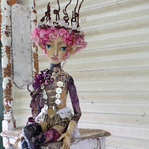May include: A whimsical doll with pink hair and a crown made of twigs and crystals sits on a white wooden chair. The doll is wearing a purple and brown dress with buttons and lace. The doll's legs are covered in purple and black patterned tights.