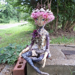 May include: A whimsical handmade doll with pink curly hair, a crown of twigs and pearls, and a purple and brown dress. The doll has a long, blue tail and is sitting on a concrete block.