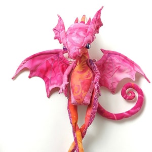 May include: A pink and orange fabric dragon with long legs and a curled tail. The dragon has wings and a patterned fabric design.