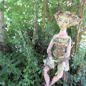 May include: A whimsical doll with a pink and white patterned dress, sitting in a forest setting. The doll has a crown of leaves and flowers on its head and is wearing a corset with lace and beads.