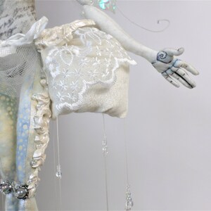 May include: A white porcelain doll with a white lace dress and a white fabric bag. The doll has a white painted face and a hand with a blue and black swirl design.