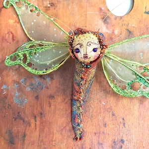 Puede incluir: Una libélula artesanal caprichosa con alas verdes y doradas y un cuerpo de tela con una cara pintada. La libélula tiene ojos azules y una boca pequeña y delicada.