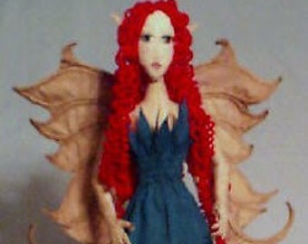 May include: A handmade fairy doll with red hair, a blue dress, and tan wings. The doll is standing on a white stand.