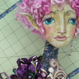 May include: A whimsical doll with pink curly hair, large blue eyes, and a painted face. The doll is wearing a purple flower, a brown and beige fabric bodice with pearl buttons, and a white lace skirt.