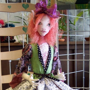May include: A handmade doll with pink hair, a green vest, a white and black polka dot shirt, and a floral skirt. The doll is sitting on a wooden chair.