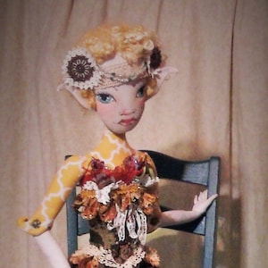 May include: A doll with blonde hair and a floral crown sits on a blue wooden chair. The doll is wearing a brown and yellow dress with a white lace trim. The doll has long, slender legs with a yellow and brown pattern.