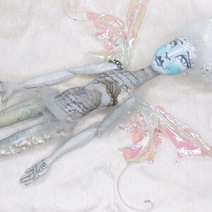 May include: A white porcelain doll with blue and white painted facial features, wearing a white dress and iridescent wings. The doll is lying on a white fabric background.