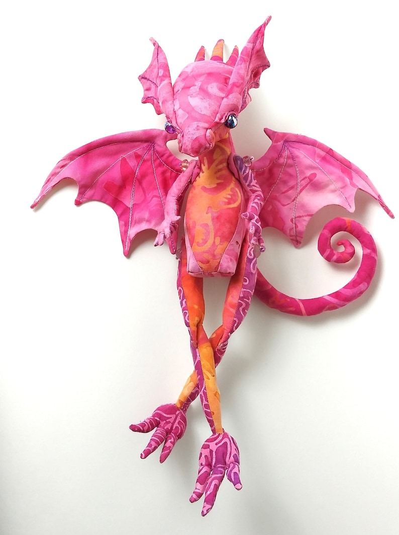 May include: A pink and orange fabric dragon with long legs and a curled tail. The dragon has wings and a patterned fabric design.