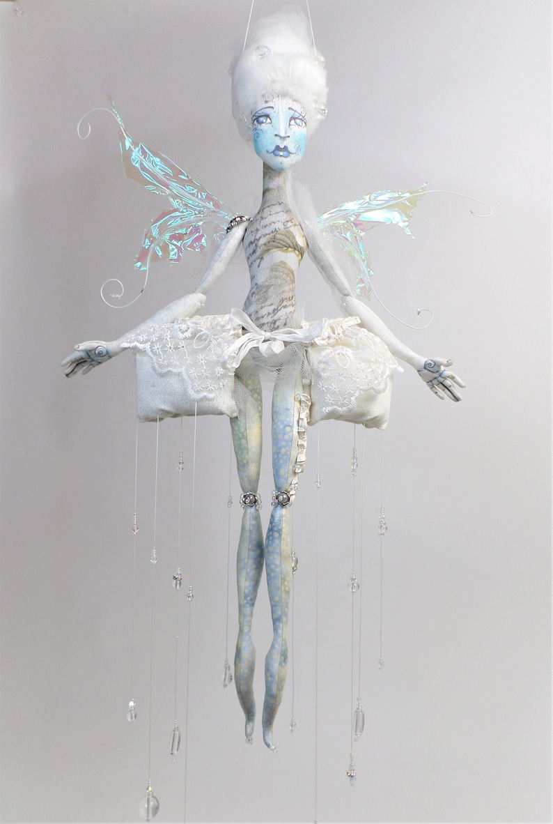 May include: A white fairy doll with iridescent wings, a white lace skirt, and blue and white patterned legs. The doll is hanging from the ceiling with strings of beads.