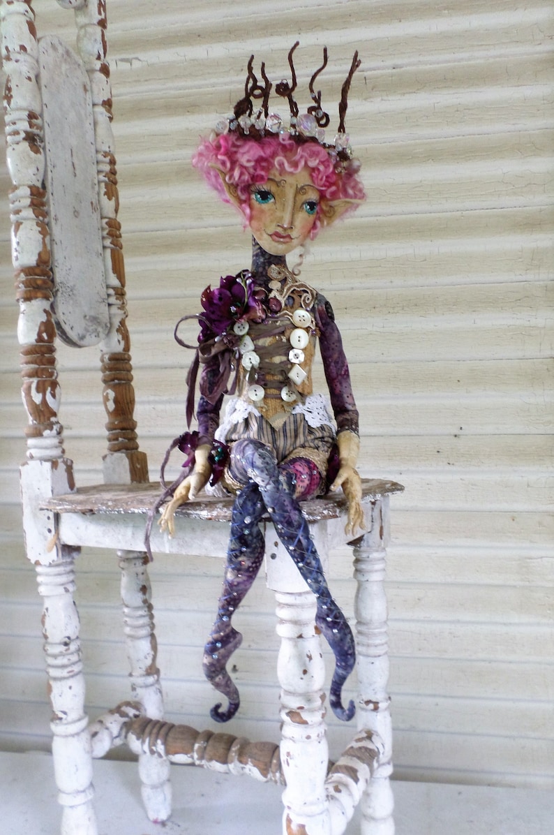 May include: A whimsical doll with pink hair and a crown sits on a white wooden chair. The doll is wearing a purple and white dress with a button and lace detail. The doll has a crown made of twigs and beads.