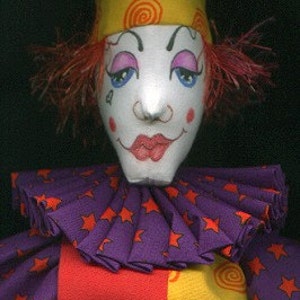 Puede incluir: Una muñeca de payaso hecha a mano con un traje morado, amarillo y rojo. La muñeca tiene una cara blanca con rasgos pintados, incluidos labios rojos, ojos azules y mejillas sonrosadas. La muñeca lleva una corona amarilla y roja.