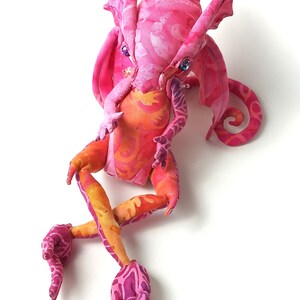 May include: A pink and orange fabric dragon with a curled tail and large wings. The dragon has a whimsical expression and is made of soft, colorful fabric.