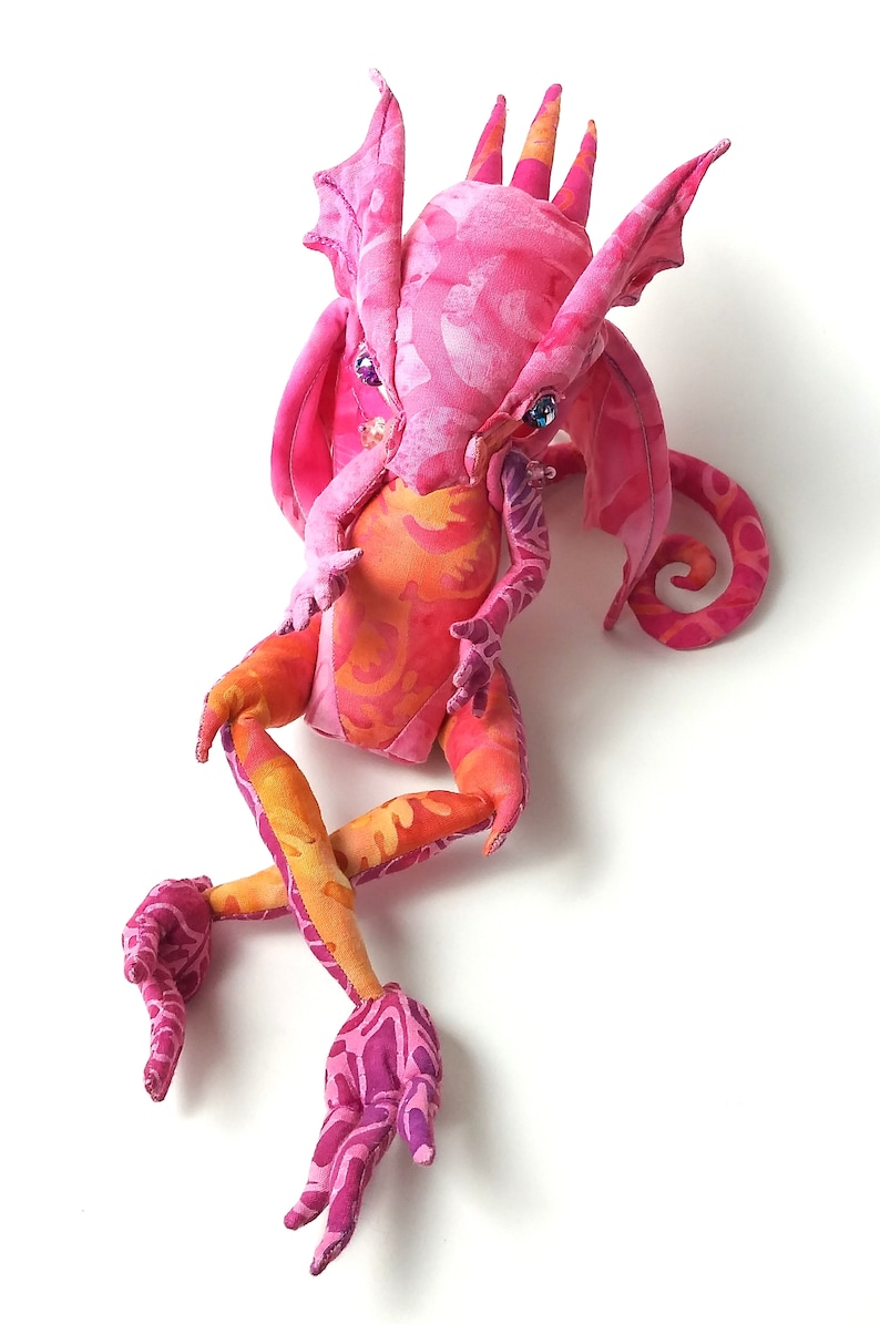 May include: A pink and orange fabric dragon with a curled tail and large wings. The dragon has a whimsical expression and is made of soft, colorful fabric.