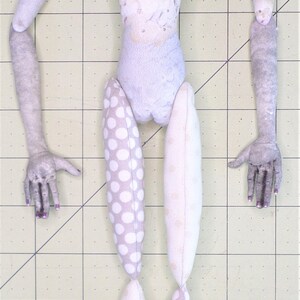 May include: A handmade doll with a white fabric body, white and gray patterned fabric legs, and gray fabric arms. The doll has a painted face with blue eyes, pink cheeks, and a pink mouth.