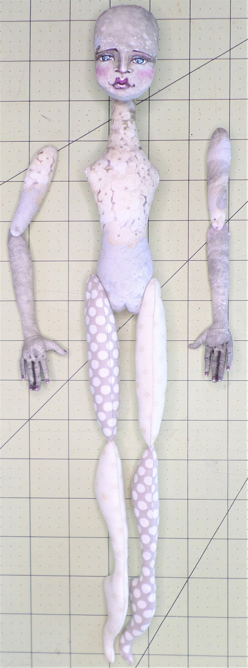 May include: A handmade doll with a white fabric body, white and gray patterned fabric legs, and gray fabric arms. The doll has a painted face with blue eyes, pink cheeks, and a pink mouth.