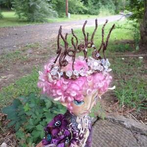 May include: A handmade doll with pink hair and a crown made of twigs and beads. The doll is wearing a purple and gold dress with a floral design.