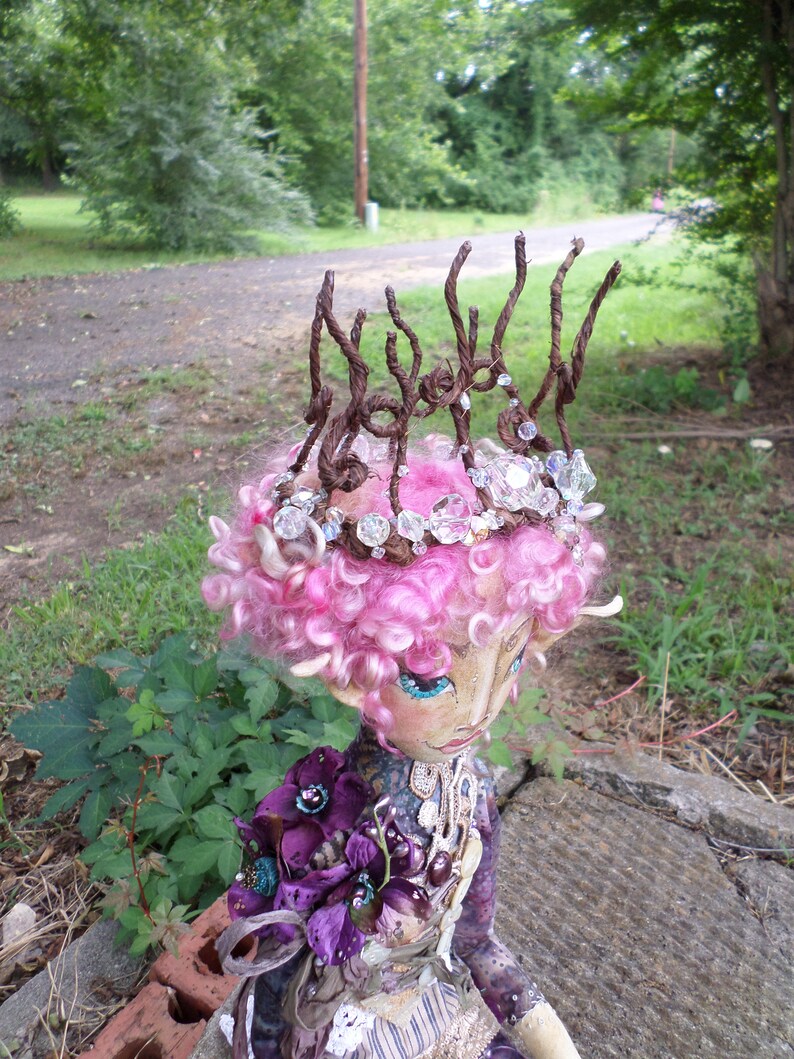 May include: A handmade doll with pink hair and a crown made of twigs and beads. The doll is wearing a purple and gold dress with a floral design.