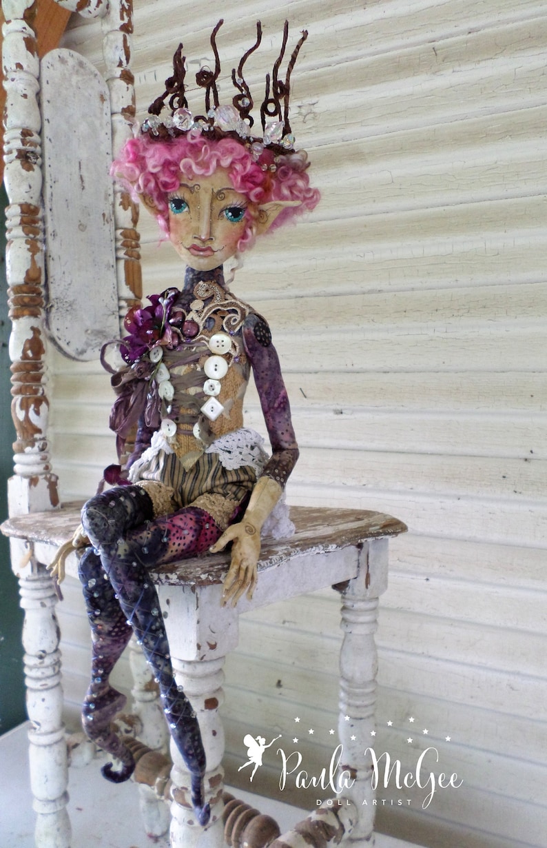 May include: A whimsical doll with pink hair and a crown made of twigs and crystals sits on a white wooden chair. The doll is wearing a purple and brown dress with buttons and lace. The doll's legs are covered in purple and black patterned tights.
