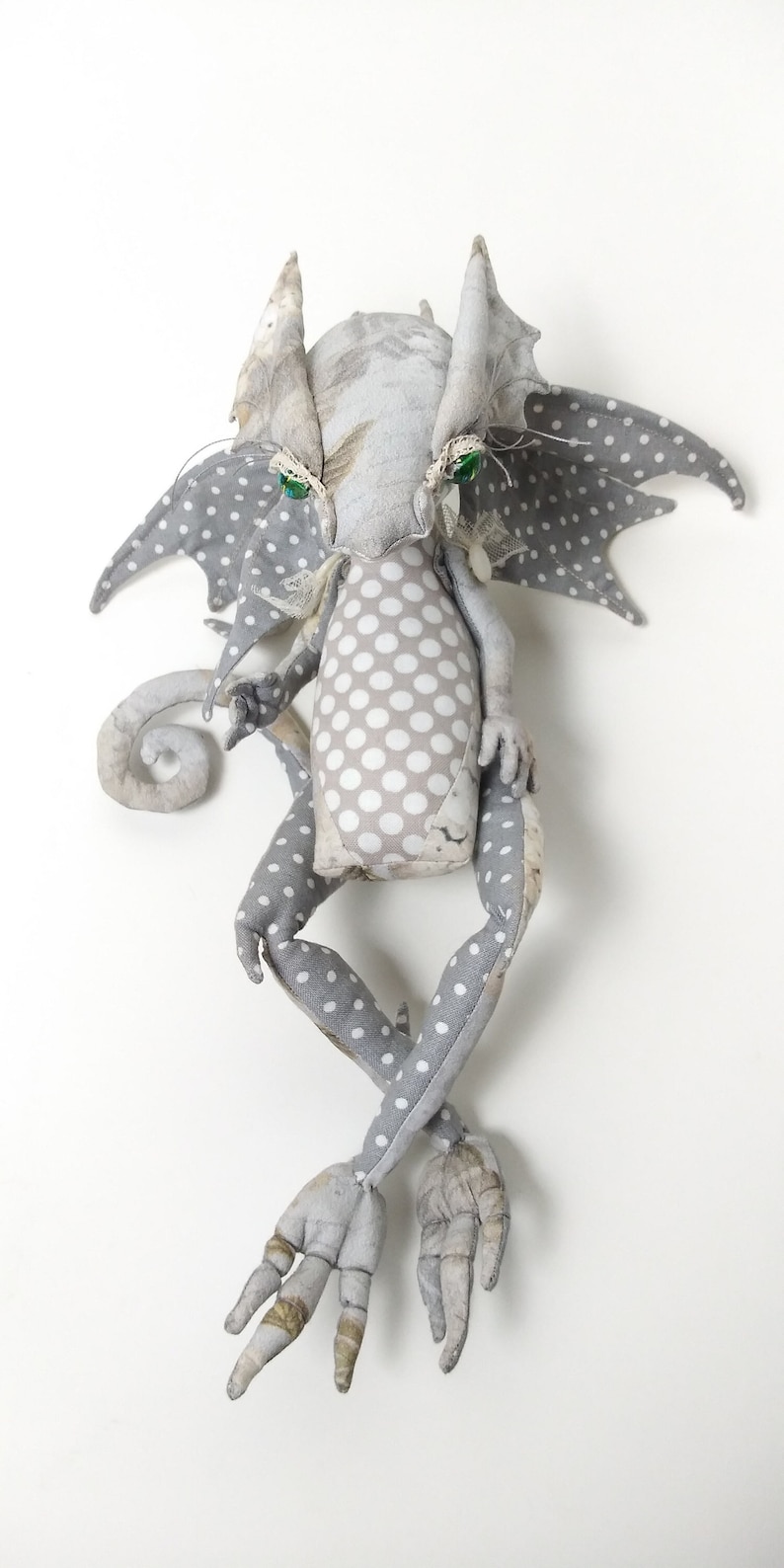 May include: A gray and white fabric dragon with green eyes and a polka dot pattern. The dragon has wings, a curled tail, and long legs with three toes on each foot.
