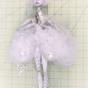May include: A white porcelain doll with a white tulle skirt and white polka dot tights. The doll has a white wig and painted facial features. The doll is holding its hands together in a prayer position.