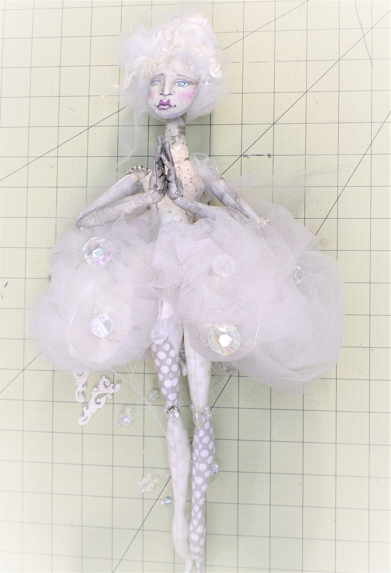 May include: A white porcelain doll with a white tulle skirt and white polka dot tights. The doll has a white wig and painted facial features. The doll is holding its hands together in a prayer position.