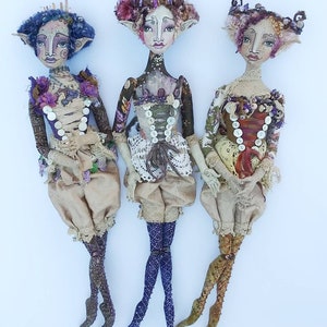 May include: Three handmade fabric dolls with whimsical features, wearing elaborate outfits with lace and buttons. The dolls have unique hairstyles and painted faces.  The dolls are standing on a white background.  "MORE DOLLS MADE BY USING WILLABELLE'S PATTERN" is written at the bottom of the image.